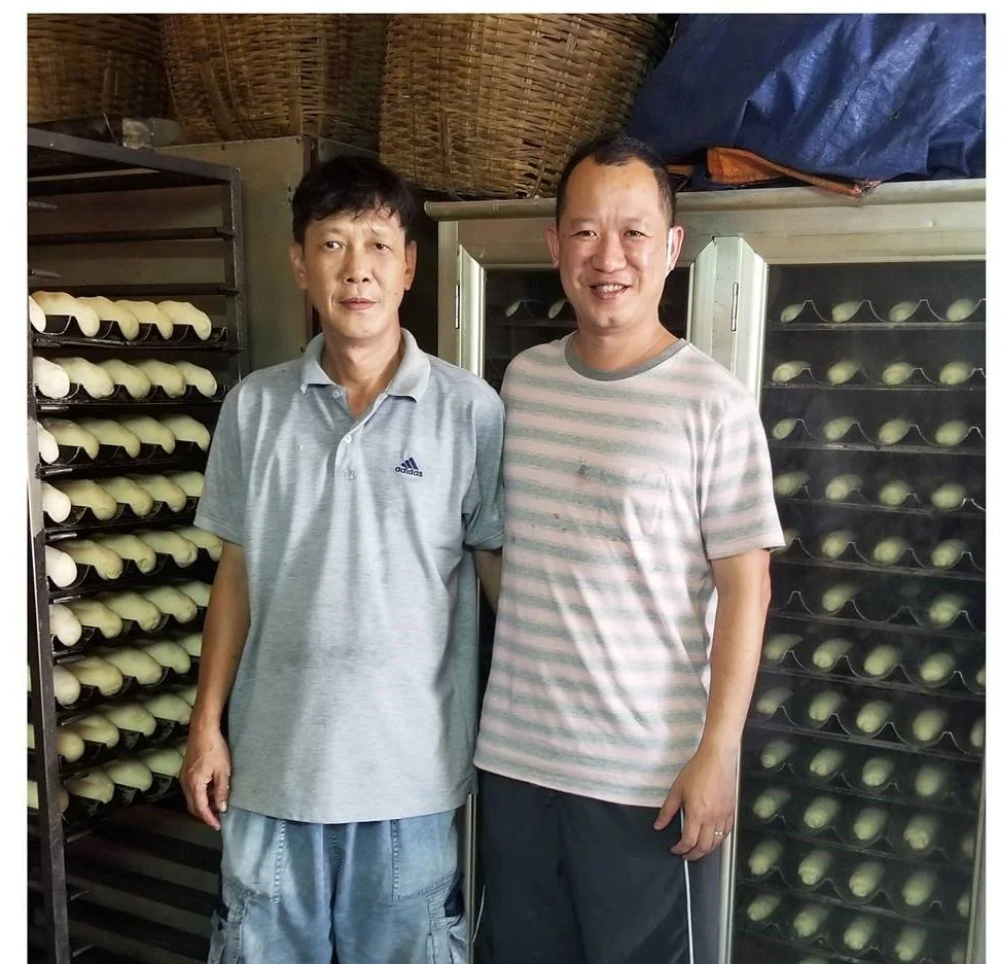 Two smiling men standing in a family kitchen surrounded by fresh-baked baguettes on trays