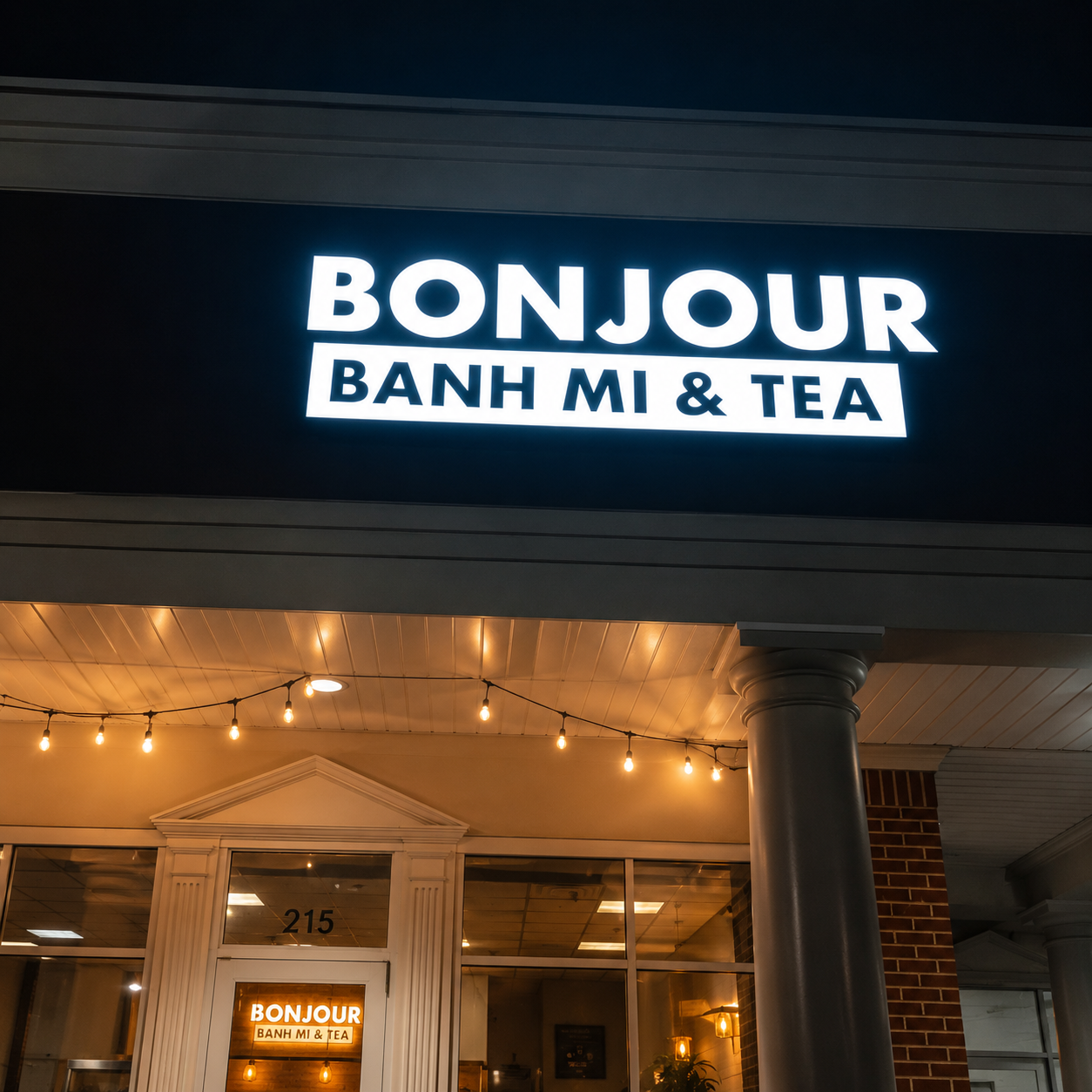 Nighttime exterior with the bright Bonjour Banh Mi and Tea sign glowing above warm string lights.